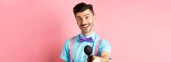 Smiling guy with moustache and bow-tie asking for comments or interview, stretch out hand with microphone, standing on pink background photo