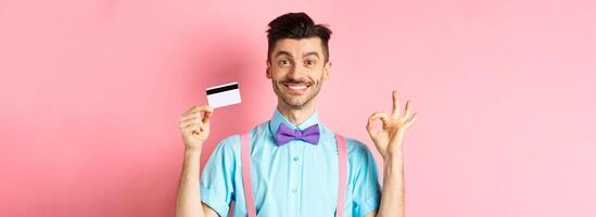 Shopping concept. Smiling handsome male shopper showing Ok sign and plastic credit card, buying something, standing satisfied on pink background photo