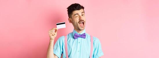 Shopping concept. Funny guy with moustache winking at camera, showing plastic credit card, recommending bank promo offer, standing on pink background photo