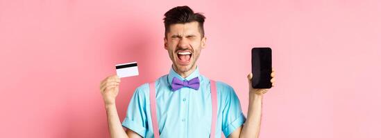 E-commerce and shopping concept. Crying man showing empty smartphone screen and plastic credit card, feeling frustrated, standing on pink background photo