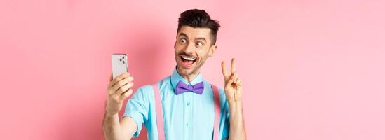 Technology concept. Funny man with moustache and bow-tie taking selfie on smartphone, showing peace sign and smiling at mobile camera, pink background photo
