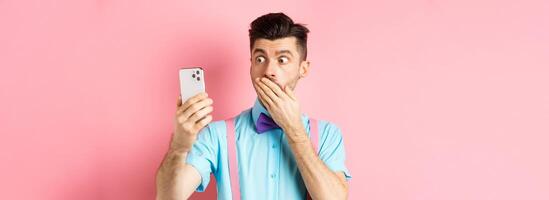 Technology concept. Silly young man looking shocked at smartphone screen while having chat, standing on pink background and cover mouth with hand photo