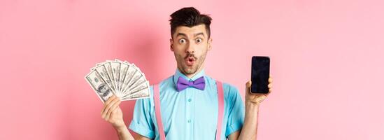 E-commerce and shopping concept. Surprised man showing blank smartphone screen and money, saying wow with amazed face, checking out online offer, pink background photo