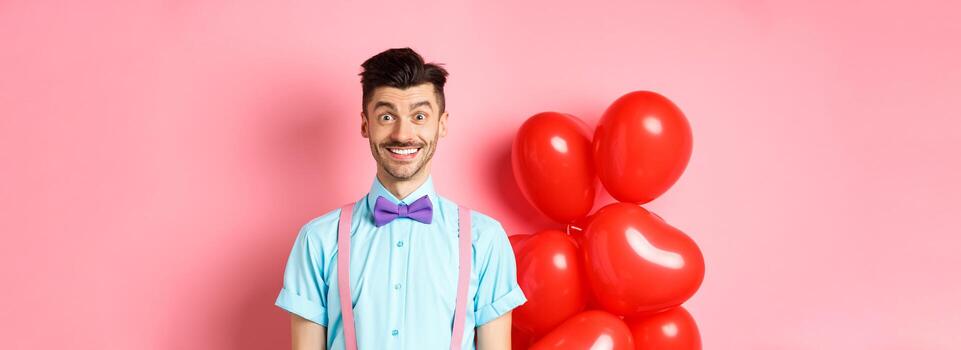 san valentin día concepto. imagen de hermoso joven hombre mirando emocionado y sorprendido, sonriente mientras en pie en romántico rosado antecedentes cerca corazón globos foto