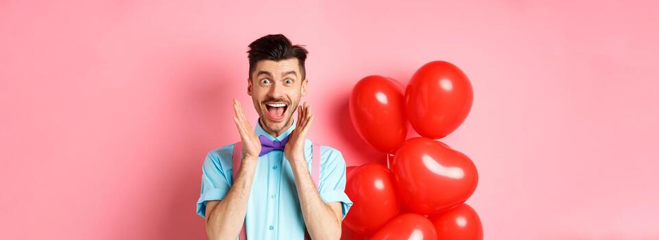 amor y romance concepto. contento hombre gritando desde fantástico noticias, gritos Guau y sonriente divertido, comprobación fuera especial oferta en san valentin, en pie cerca rojo corazones globos foto