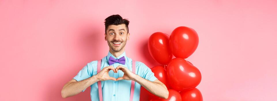 san valentin día concepto. romántico chico en corbata de moño, demostración corazón gesto y decir yo amor usted con esperanzado sonrisa, en pie en rosado antecedentes cerca globos foto