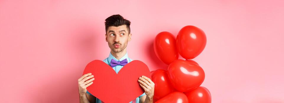 san valentin día concepto. encantador joven hombre en corbata de moño esperando para alma gemela con grande rojo corazón separar, mirando izquierda y sueño de amar, rosado antecedentes foto
