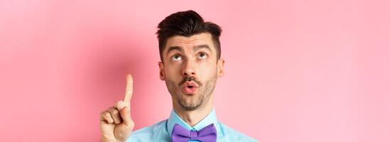 Intrigued young man in moustache, pointing finger and looking up at something interesting, checking out advertisement, standing over pink background photo