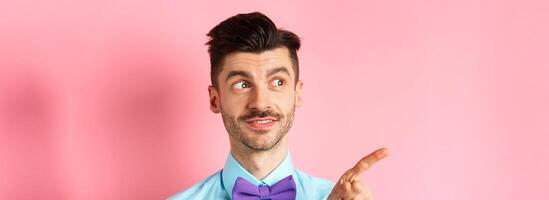 Close-up of caucasian male model with moustache and bow-tie, looking and pointing left with intrigued smile, showing promo offer, standing in bow-tie on pink background photo
