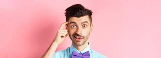 Close-up of confused guy in bow-tie, scratching head and looking puzzled, cant understand something, standing in bow-tie on pink background photo