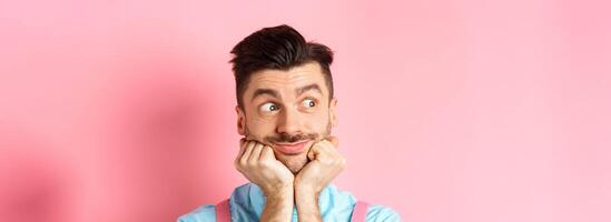 Close up of cute guy dreaming of something, looking aside and imaging things, standing on pink background, leaning face on hands, pink background photo