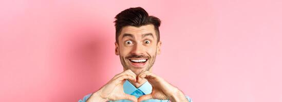 Valentines day concept. Close up of romantic guy looking happy, smiling and showing heart gesture, standing on pink background photo
