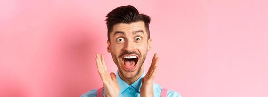 Close-up of happy caucasian guy screaming of joy and surprise, checking out advertisement, seeing something cool, standing over pink background photo