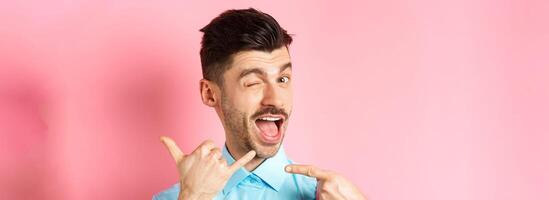 Cheeky and funny guy showing phone gesture, asking to call him and winking, telling dial his number, standing over pink background photo