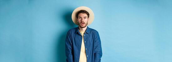 Young man tourist in summer hat drop jaw, staring at something shocking, standing on blue background photo
