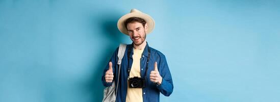 Handsome caucasian tourist in straw hat, holding backpack and photo camera, showing thumbs up, recommending travel agency or hotel resort, blue background