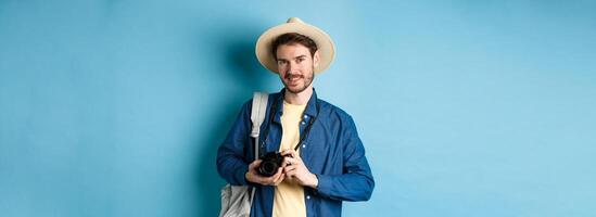 Handsome guy going on summer vacation, backpacking on holiday. Tourist with straw hat and camera smiling happy, standing on blue background photo