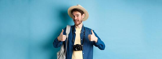 Happy smiling tourist say yes and show thumbs up, going on summer vacation, holding backpack and photo camera, blue background