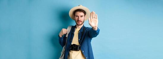 Serious male tourist warn you, stretch out hand to show stop gesture. Man on vacation forbid something, refusing or prohibiting action, standing with backpack on blue background photo