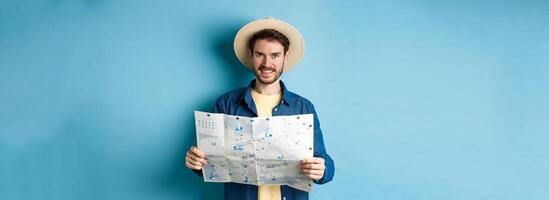 Happy and positive guy on vacation, looking at camera and holding map, smiling excited, going on summer travel, standing on blue background photo