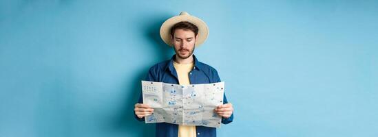 Handsome tourist with summer hat reading map, looking for sightseeing on vacation, standing on blue background photo
