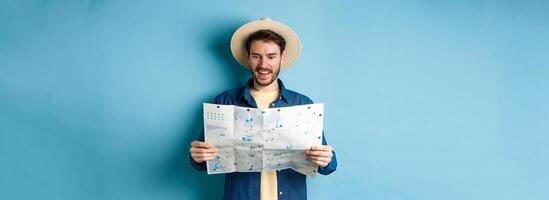 Cheerful tourist in summer hat planning travel route on vacation, looking at sighseeing map and smiling excited, standing on blue background photo