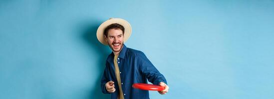 Happy man laughing and playing frisbee, throwing at friend and smiling, standing in summer hat on blue background photo