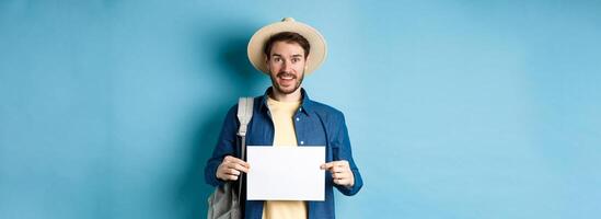 Cheerful tourist in summer hat, showing empty piece of paper and smiling, hitchhiking with backpack, standing on blue background photo