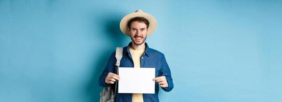 Happy tourist with backpack showing empty piece of paper, smiling at camera, standing on blue background. Concept of summer holiday and vacation photo