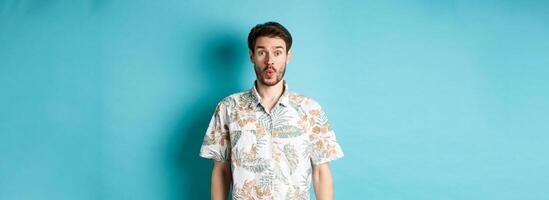 Summer holiday. Surprised tourist say wow and staring at camera, checking out awesome promo, standing in hawaiian shirt on blue background photo