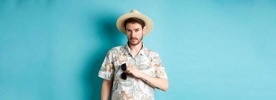 Handsome tourist in hawaiian shirt and summer hat, put sunglasses in pocket, going on vacation, standing against blue background photo
