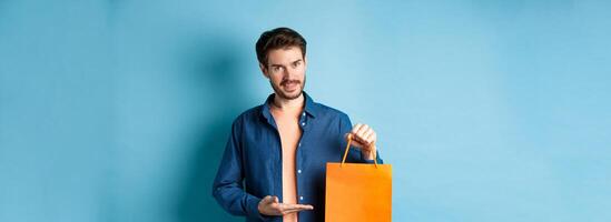 Handsome caucasian man showing orange shopping bag and smiling, purchased gift, standing on blue background photo