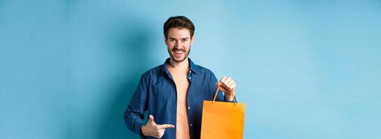 Smiling caucasian man pointing finger at orange shopping bag with bought items, standing on blue background photo