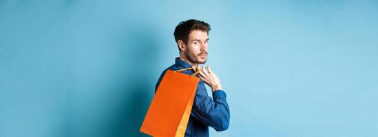 Rear view of happy man holding shopping back on shoulder and looking behind, standing on blue background photo
