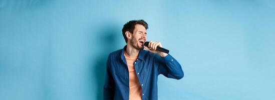 Carefree guy singing karaoke with microphone, perform a song, standing on blue background photo