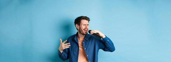 Romantic guy singing song in microphone and gesture, singer playing karaoke, standing on blue background photo