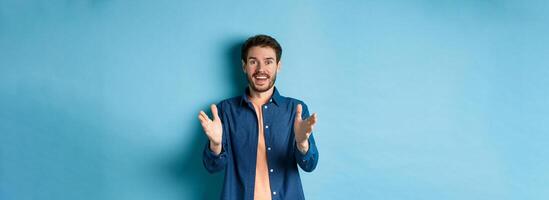 Cheerful young man pointing hands at camera and smiling, feeling glad for you, praising nice work, standing on blue background photo