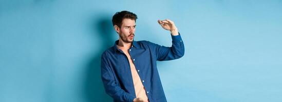 Impressed caucasian guy staring at empty space with big object, shaping large size, standing on blue background photo