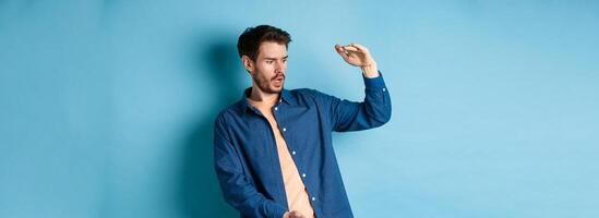Shocked young man looking at empty space and showing big size object, standing on blue background photo