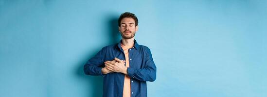 Romantic and nostalgic guy holding hands on heart with eyes closed, keep warm memories, standing on blue background photo