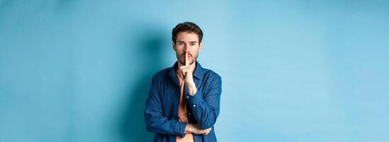 Modern guy shushing at camera, press finger to lips and telling a secret, making taboo sign, standing on blue background photo