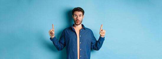 Surprised young man say wow, checking out advertisement, pointing fingers up and looking impressed, standing on blue background photo