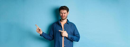 Disappointed young man frowning and pointing fingers right at empty space, looking upset and displeased, complaining, standing on blue background photo