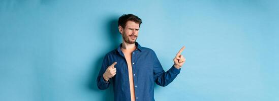 Skeptical young man pointing and looking left with disappointment, cringe from something bad, standing upset on blue background photo