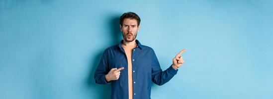 Shocked and offended young man frowning and looking hurt, pointing fingers left at empty space, complaining on something, standing on blue background photo