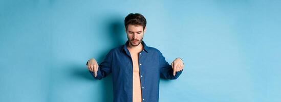 Image of modern guy in casual clothes, looking and pointing fingers down at empty space, reading logo, standing on blue background photo