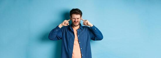 Annoyed young man block sound, shut ears with fingers and grimacing, disturbed by loud music, standing on blue background photo