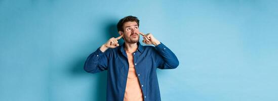 Annoyed young man block sound, shut ears with fingers and grimacing, disturbed by loud neighbours, looking up irritated, standing on blue background photo