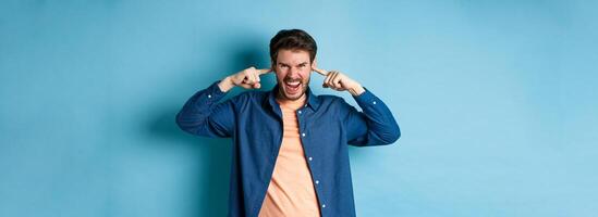 Angry young man shut ears to block annoying sound and shouting at camera, frowning and looking furious, standing on blue background photo