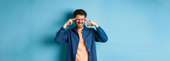 Man feeling sick, touching head and grimacing from painful migraine, having headache or feel dizzy, standing on blue background photo